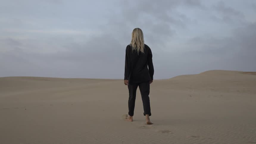 Woman walks barefoot across the dunes under a cloudy quiet sky