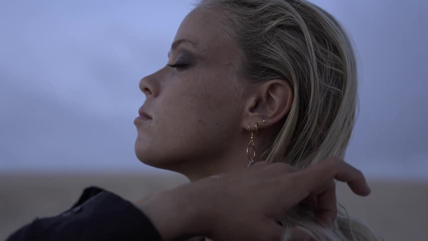 Send a close side view shows a woman with long wet blonde hair and dramatic eye makeup wearing an earring as a hand is briefly seen pulling her hair back against a pale gray and blue sky