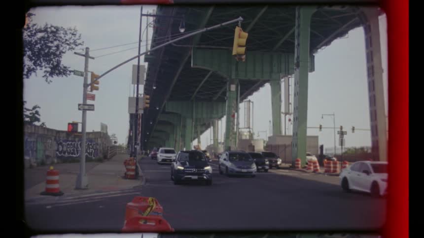 Cars drive beneath an overhead bridge on a sunny day casting moving shadows across the bright pavement