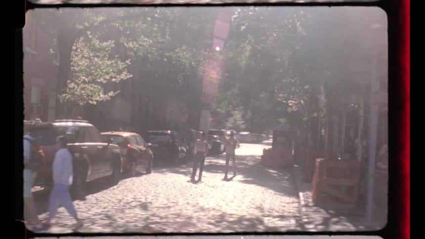 The video is shot with a film frame border and appears to be a firstperson view from a moving vehicle traveling through a sunny city street the footage shows glimpses of brownstonestyle buildings parked cars pedestrians and street signs