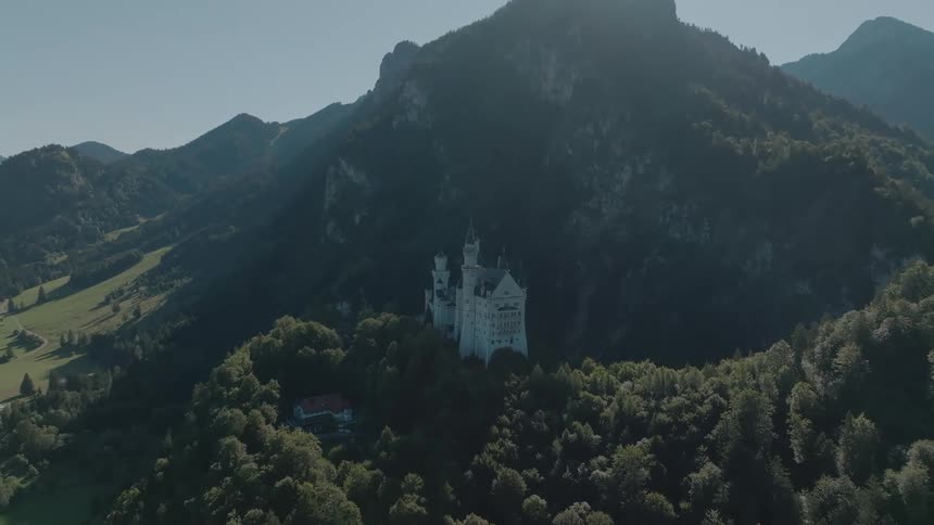 An aerial view shows a white castle situated amidst green trees on a hill framed by massive dark wooded mountains under a sunny sky