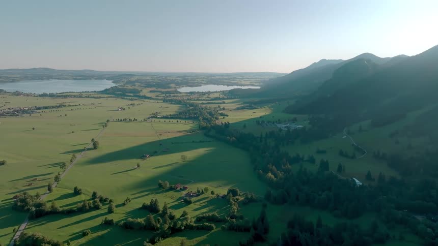 An aerial shot shows a wide green valley containing scattered houses and extending toward distant lakes bordered on the right by a large mountain range under a clear sky