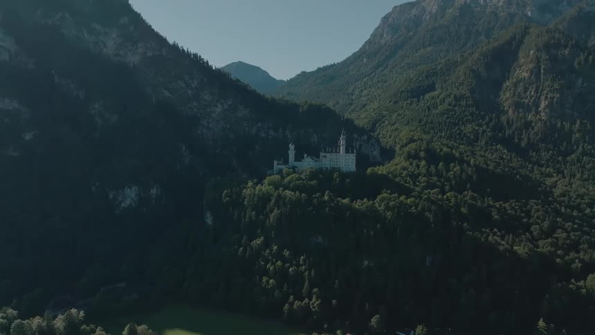 A castle stands atop a hill surrounded by mountains and lush green vegetation under the bright midday sun