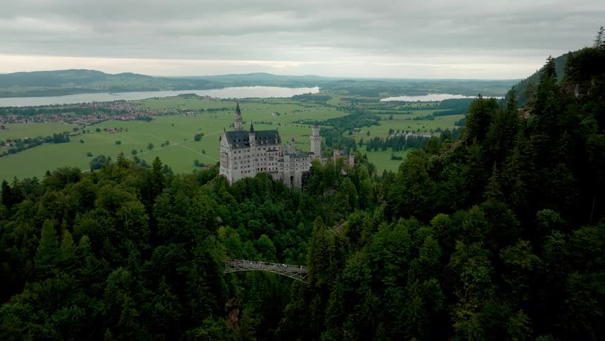 An aerial shot looks down onto a castle situated on a heavily forested hill with a wooden bridge visible below and a broad green valley with lakes and a town stretching out to the horizon under a cloudy sky