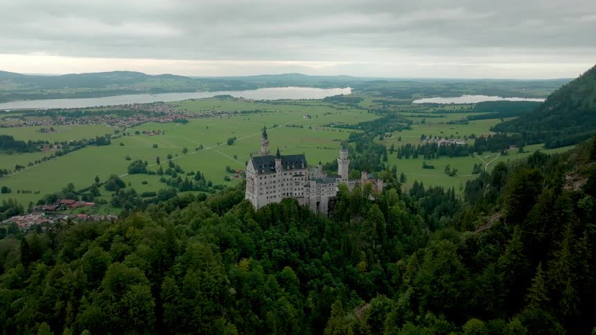Aerial view of a fairytale castle on a forested hill overlooking fields and a lake