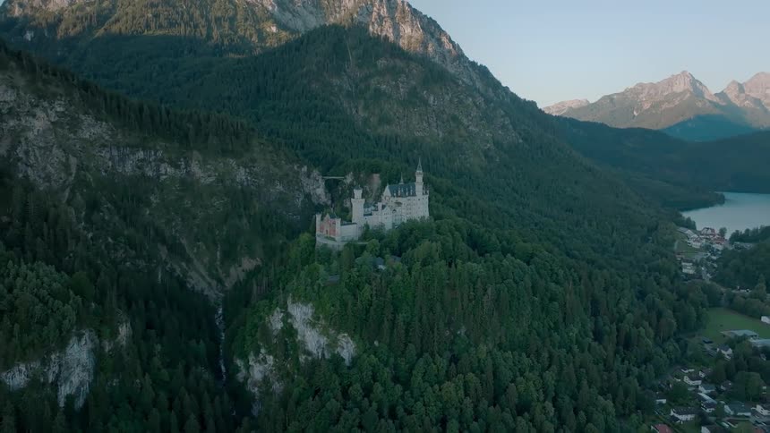 Aerial view of a castle on a forested ridge with mountains and a distant lake