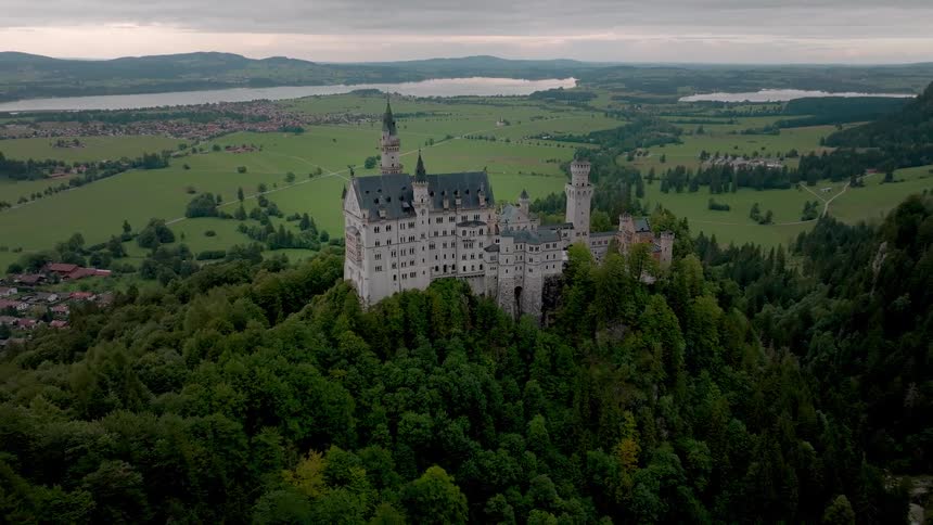 A castle stands atop a hill surrounded by trees in a quiet rural area overlooking a small town nestled in the lower land below