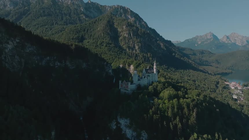 An aerial view shows a large castle perched on a hill surrounded by dense green forest situated in a valley between steep rockfaced mountains
