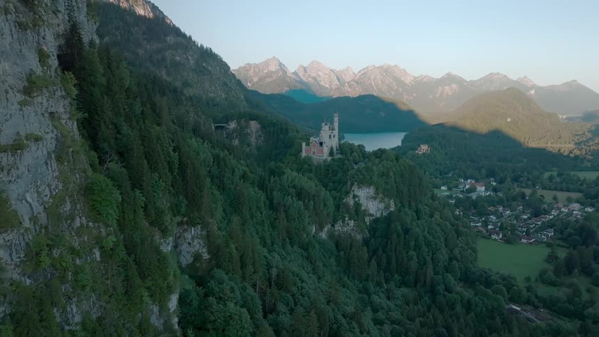 A castle perched on a hill encircled by trees and mountains overlooks a town nestled below at midday