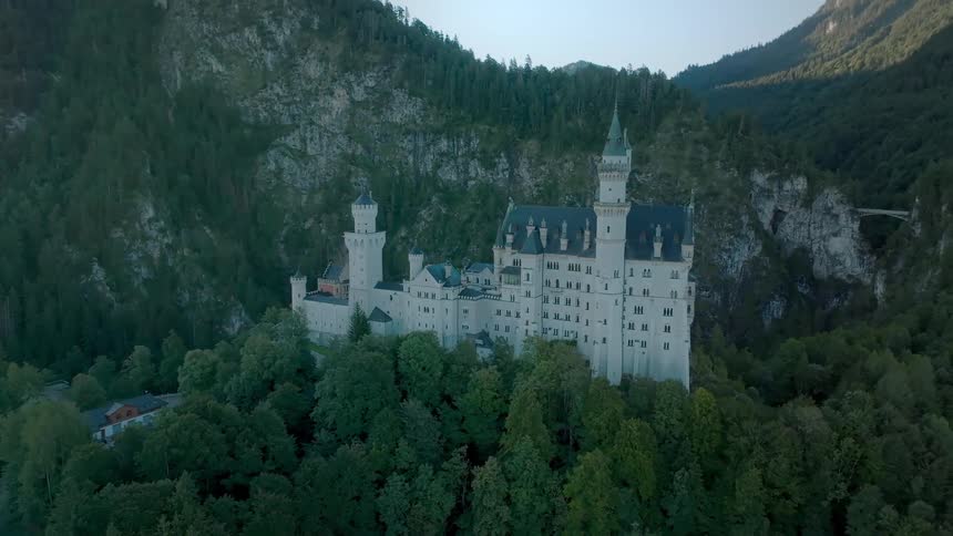 A sprawling lightcolored castle with multiple towers sits atop a forested hill surrounded by dark green trees and framed by steep rugged mountains