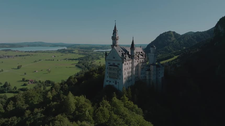 Neogothic castle nestled among green hills overlooking a valley and lake in the distance