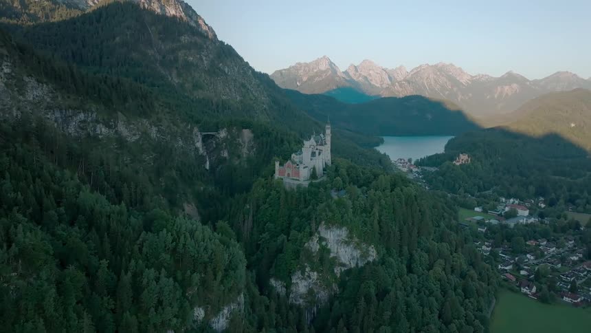 A castle perched on a hill encircled by trees and mountains overlooks a town nestled below at midday