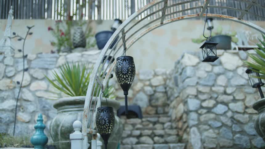 Two decorative black lanterns hang from a curved metal archway in the foreground with a stone staircase and various potted plants ascending a terraced stone retaining wall in the background