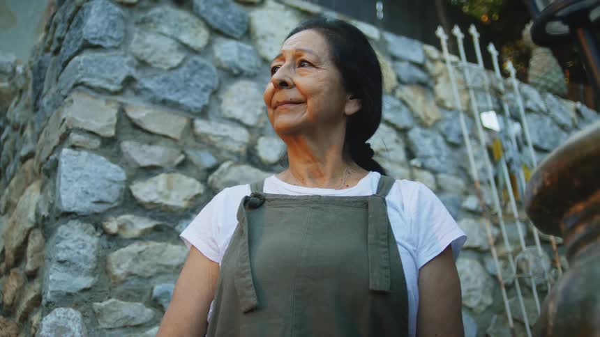 An older woman wearing a white shirt and green overalls stands outdoors before a stone wall looking upward with a contemplative expression and with a warm light on her face