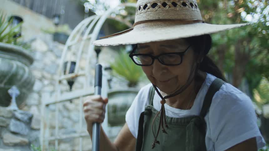 An elderly woman wearing a hat sweeps the outdoor area of a building under the bright midday sun