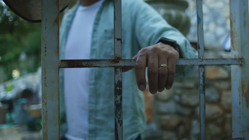 Man rests his hand on a rusty metal gate in an outdoor setting