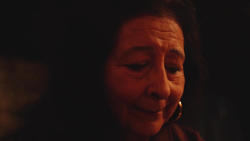 In a dimly lit close up an older woman with a gentle emotional expression looks down and smiles