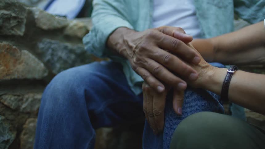 Two people sit close together outdoors with one hand resting on the other persons hand touching softly on their fingers in front of a stone wall