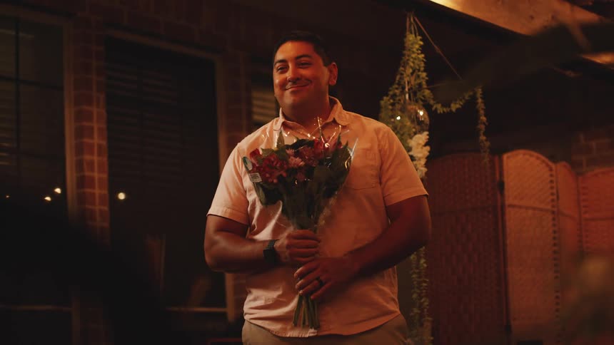 Man smiles while holding a bouquet of flowers in a warmly lit setting