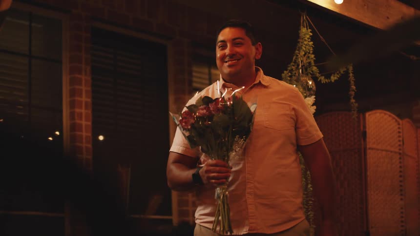 The video is a medium shot of a man standing outdoors on a patio at night smiling broadly and holding a bouquet of dark pink and red flowers the area behind him is decorated with warm string lights and hanging greenery