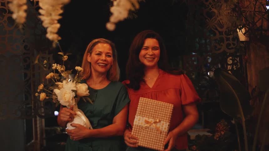 Two women smile holding flowers and a gift during an evening celebration