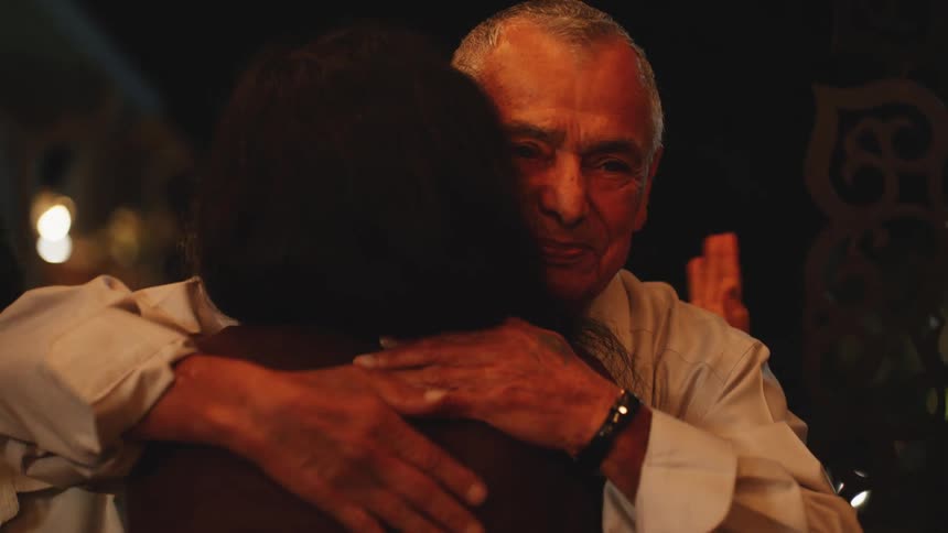 A close view shows an older man wearing a white collared shirt happily embracing a woman with her back to the camera in a dimly lit indoor setting