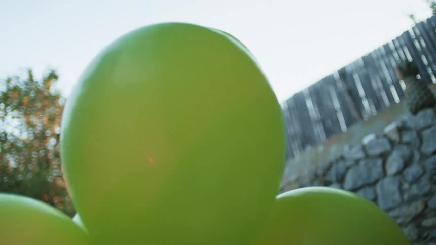 A cluster of large green balloons sits outdoors against a blue sky and stone wall