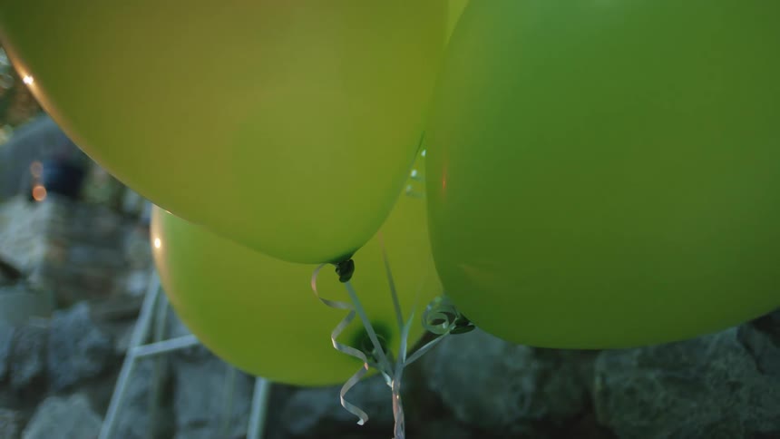 A close low angle view shows a cluster of bright shiny lime green balloons floating against a background of dark stone wall and metal trellis