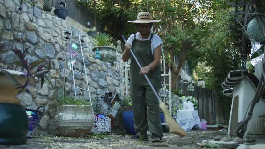 A woman wearing a hat and overalls sweeps an outdoor stoneanddirt path with a straw broom working on the ground in front of a tall tiered stone retaining wall decorated with various pots and plants