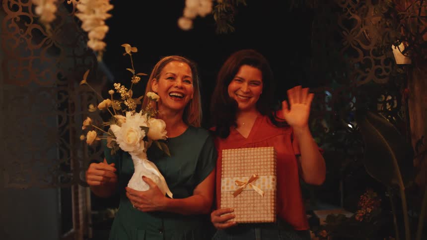 Two young women stand side by side at nightone holding a gift bag the other a flower plantas they wave warmly to someone in the distance under the glow of streetlights
