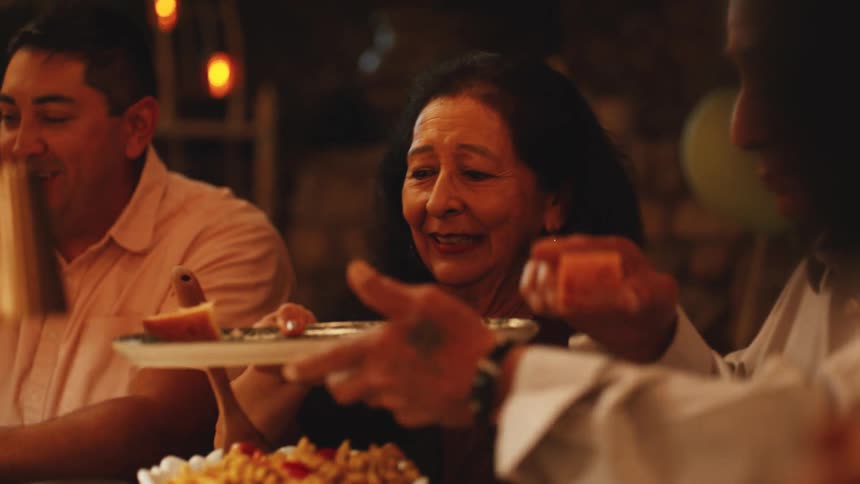A group of people sit around a table in a dimly lit room where one person holds out a plate of sliced fruit toward an older woman who smiles while another man reaches for his glass
