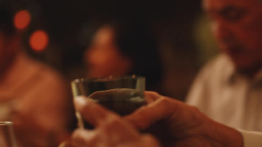 A dark liquid is poured from a pitcher into a glass held by a persons hand while the action takes place at a table with other people visible in the dark background