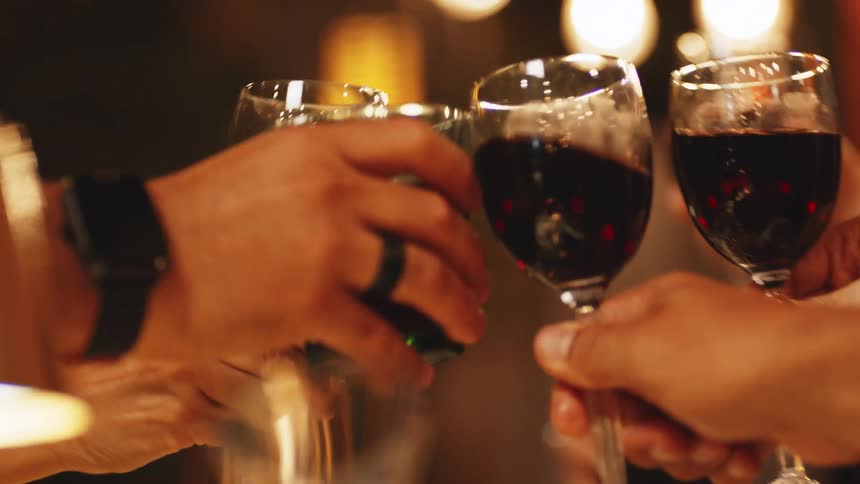 A group of people clink glasses and toast each other with red wine at a warmly lit table