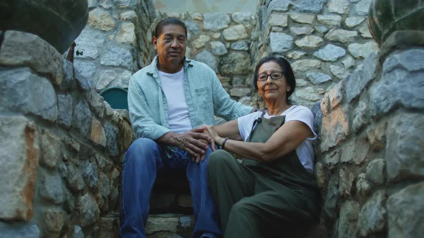 A man and a woman sit together on a stone staircase framed by stacked stone walls resting their hands on their knees while looking directly at the camera