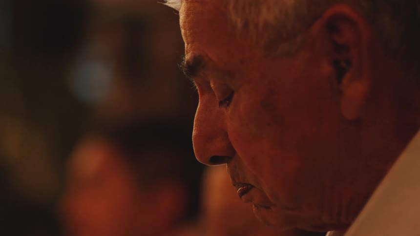 A closeup profile shot of an older mans with white hair eating against a warm blurred background with other people out of focus