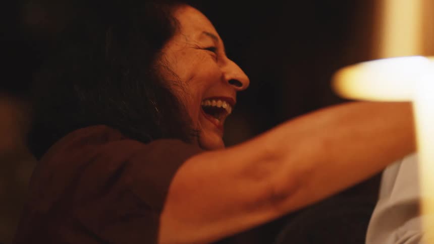 An elderly woman throws her head back and laughs joyfully in a warmly lit intimate setting