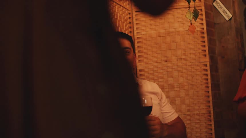 A close dimly lit view shows a middle aged man smiling while holding a glass of red wine and an older woman smiling next to him at an outdoor party with a bamboo accent wall watching people dance