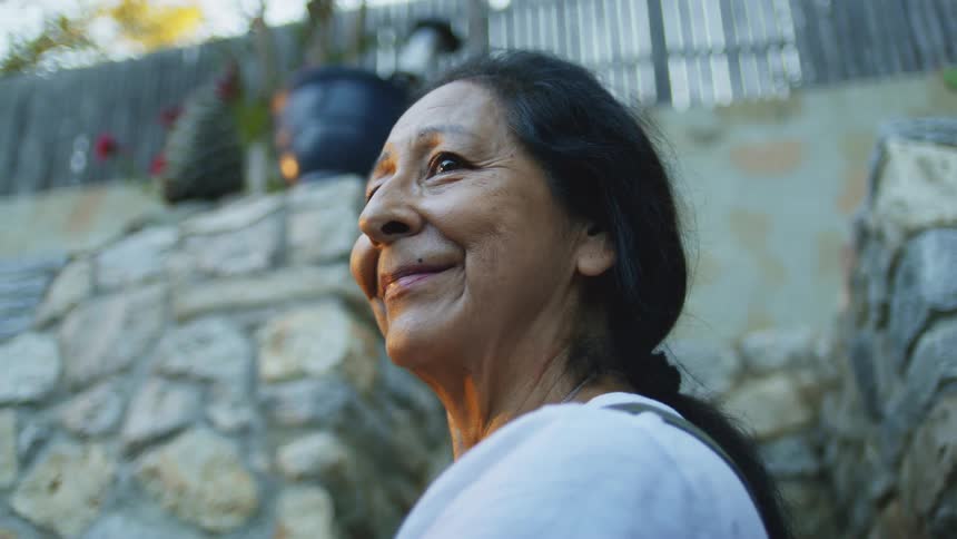 An elderly woman with a warm smile gazes upwards against a backdrop of stone and greenery