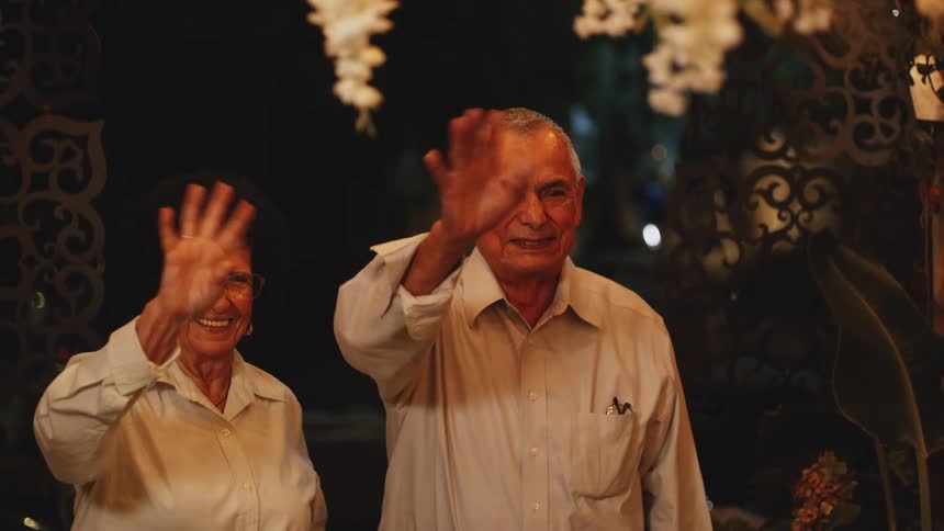 A smiling elderly couple stands together at night happily waving to someone off camera