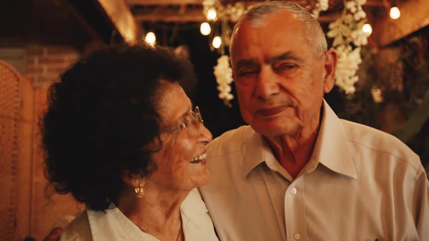 An affectionate elderly couple smiles at each other and the man leans in for a kiss under warm party lights