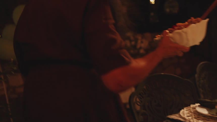 A smiling man and woman bring bowls of food to a warmly lit dinner table for an evening gathering