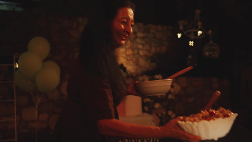 A middle aged man wearing a blue collared shirt and a middle aged woman with long dark hair wearing a brown blouse are setting out food for a dinner party at a table decorated with candles and a lamp on a dark patio with a stone wall