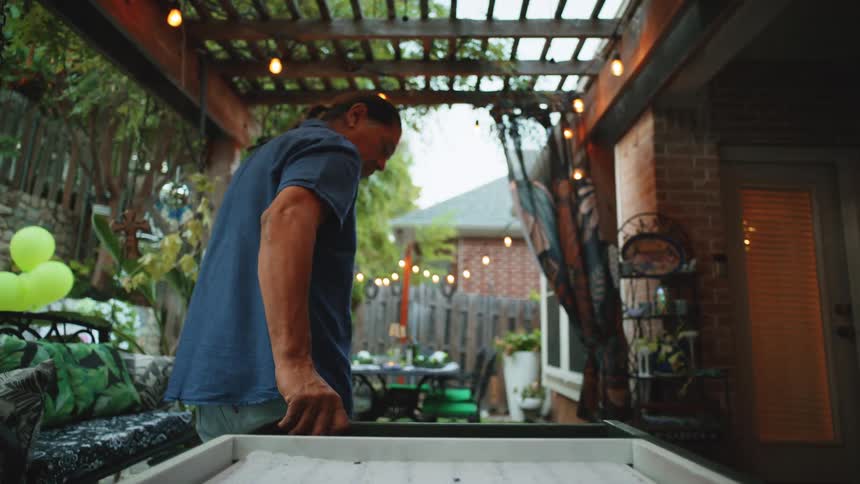 A man in a blue shirt with his hair pulled back is standing over a white tray on a patio under a wooden pergola decorated with lights and flowers overlooking a green backyard