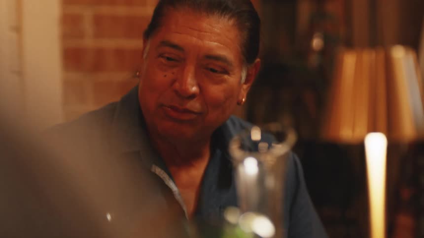 A closeup shot of a mans face at a dinner table shows him smiling and looking forward then getting a wine glass and putting at the table