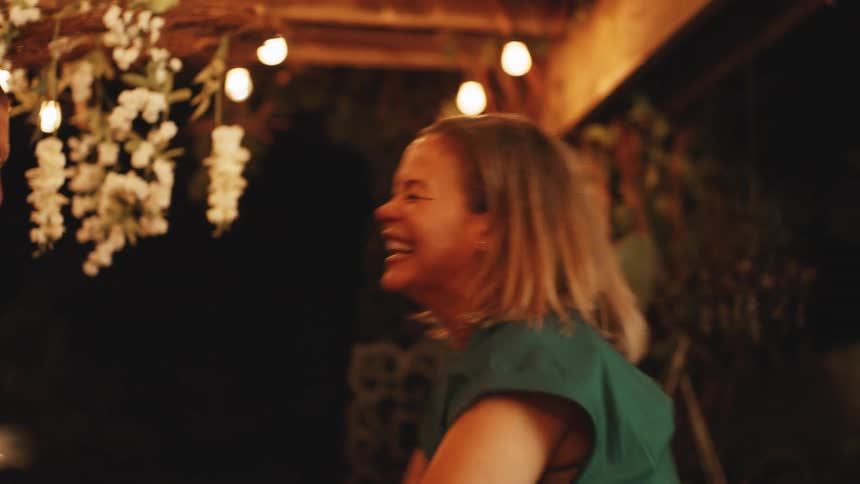 A camera shot shows a man and a woman holding hands while dancing and laughing with overhead string lights and white flower decorations visible in the background of the dark patio