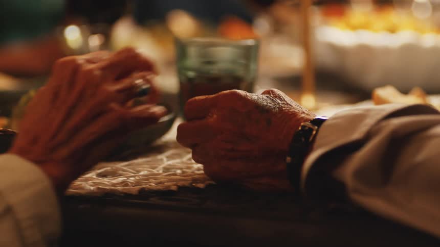 Elderly hands clasp together over a dinner table lit by candlelight