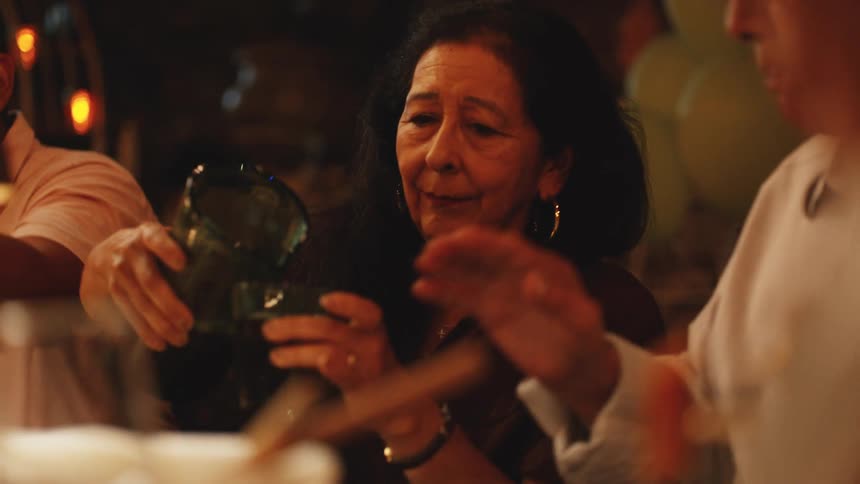 An elderly woman pouring a cup of drink from a jug while seated at an indoor family dinner at night