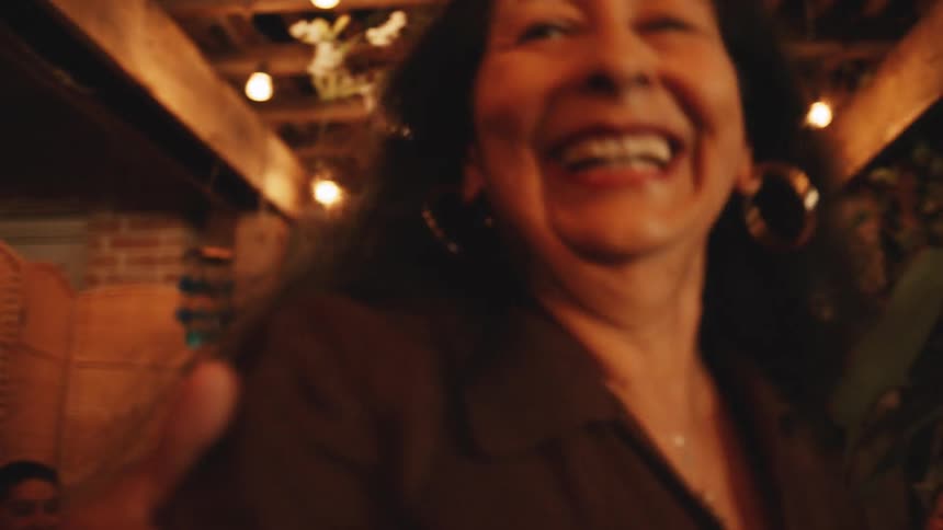A middle aged man wearing a dark short sleeved collared shirt and a woman with long dark hair laugh while happily dancing together at an evening party illuminated by string lights and surrounded by smiling guests and white floral decorations