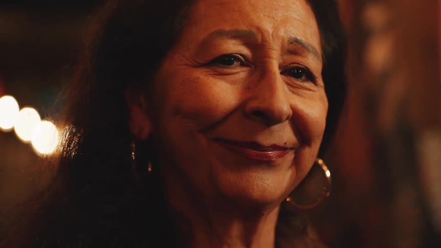 A close view shows a smiling older woman with long dark hair and large gold hoop earrings illuminated by a row of out of focus string lights in the background