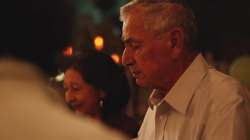 An elderly man speaks while seated next to his wife at a warmly lit gathering